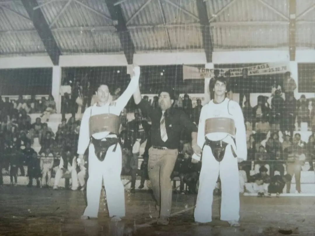 Foto antiga em preto e branco de um árbitro levantando o braço do vencedor de uma luta de Taekwondo num ginásio lotado