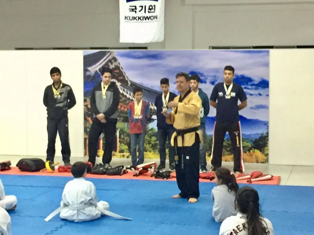 Mestre Paulo Ribeiro de dobok dourado fala com crianças sentadas no tatame, com jovens medalhistas em pé ao fundo, diante de painel coreano e bandeira do Kukkiwon