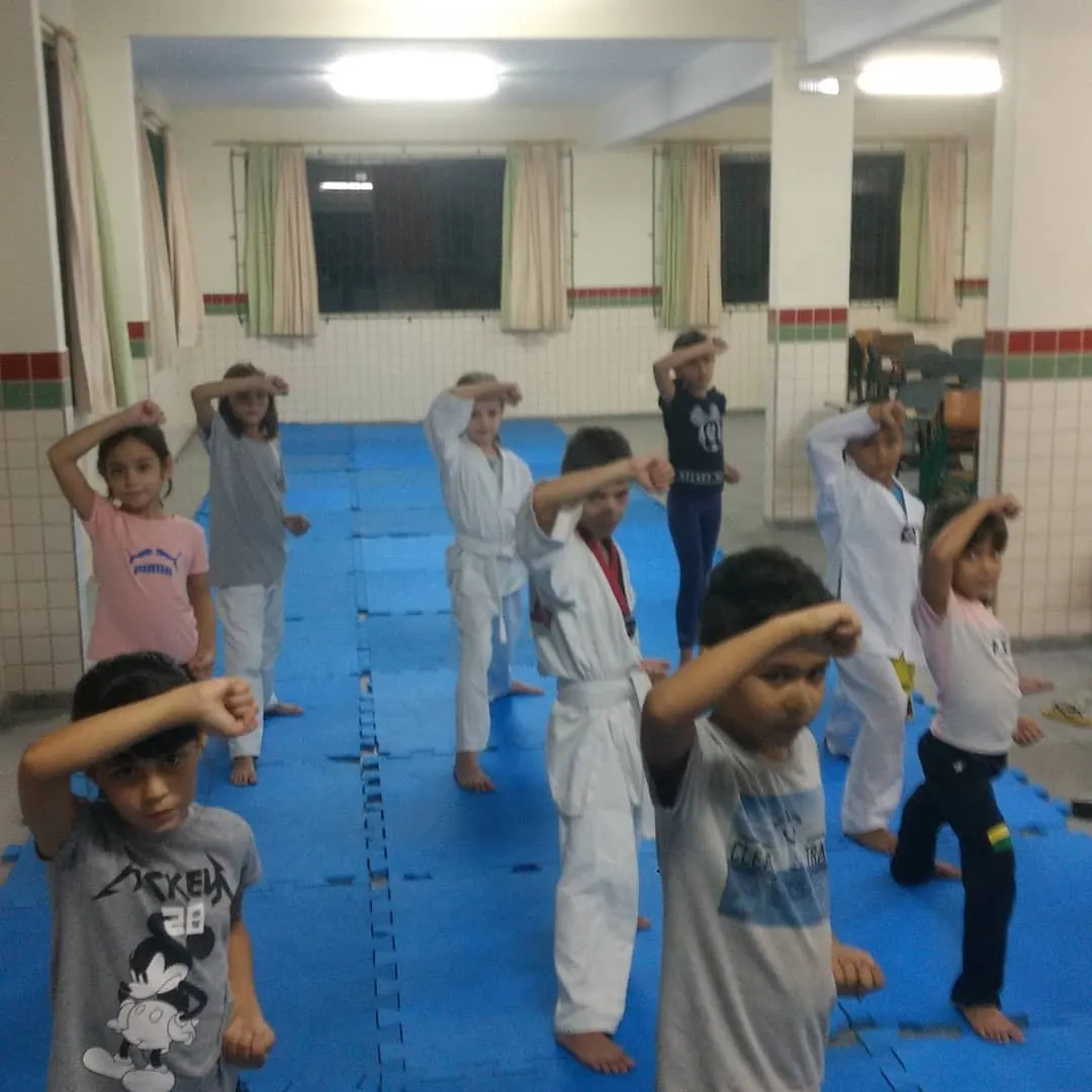 Crianças treinando defesa de Taekwondo com o braço levantado sobre o tatame azul numa sala de escola