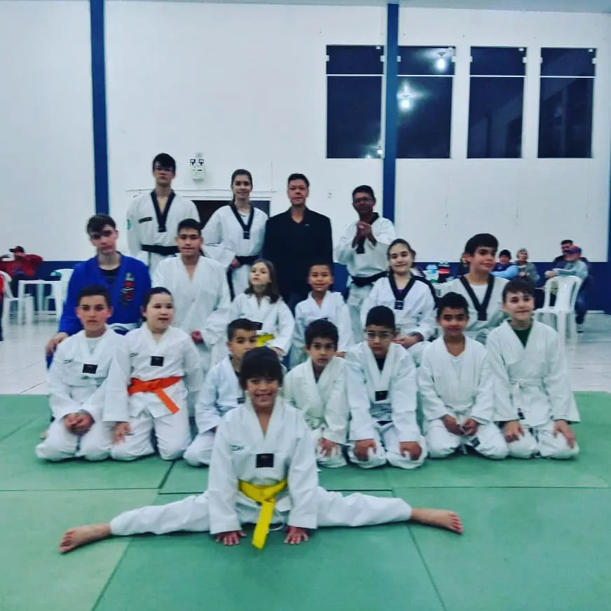Turma infantil de Taekwondo de dobok branco reunida no tatame verde com o Mestre e os instrutores ao fundo