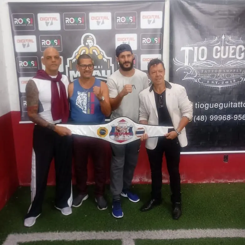Mestre Paulo Ribeiro e outros três homens segurando um cinturão de campeão de Muaythai diante de painel de patrocinadores