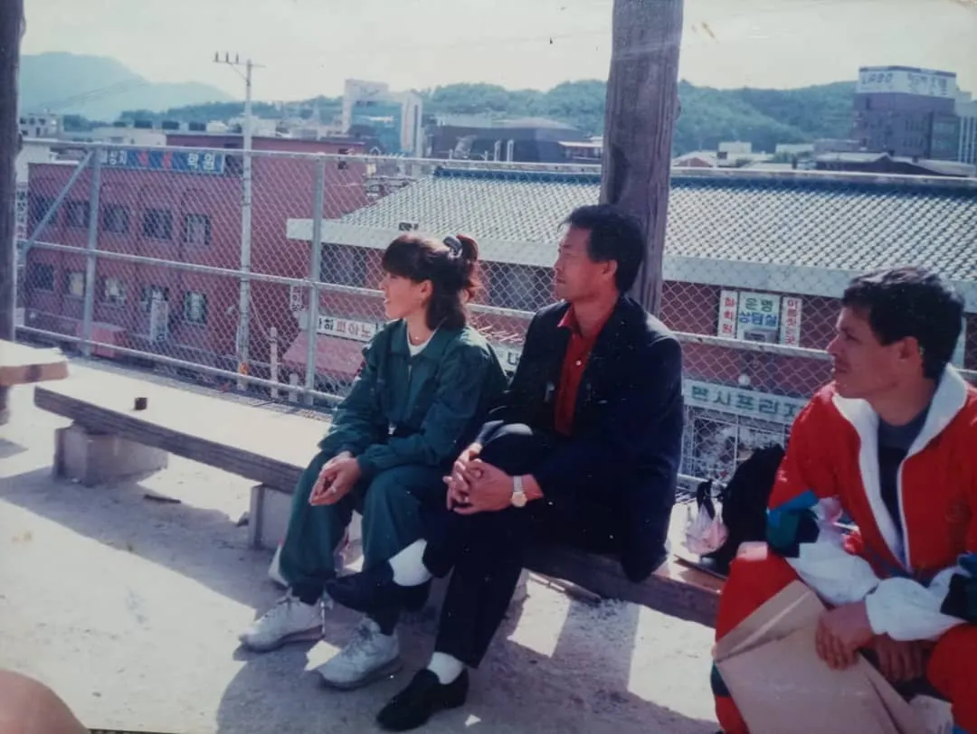 Foto antiga de três pessoas sentadas num banco ao ar livre, com prédios e placas em coreano ao fundo