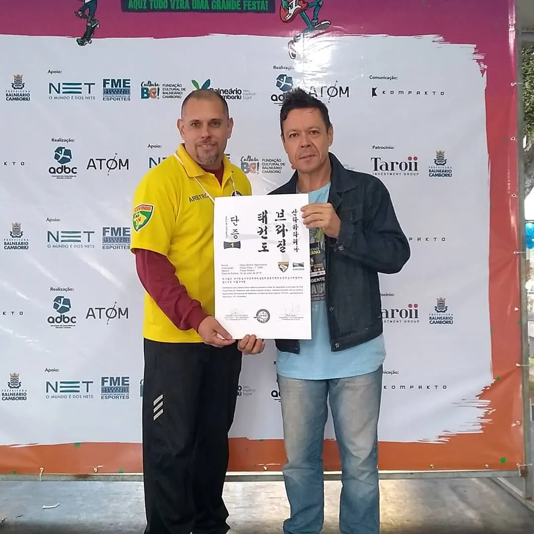 Mestre Paulo Ribeiro segurando com um aluno adulto o certificado de faixa preta, diante do painel de patrocinadores de Balneário Camboriú