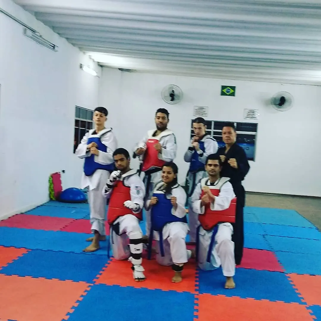 Seis atletas adultos com coletes de luta e o Mestre de dobok preto, em guarda no tatame da academia