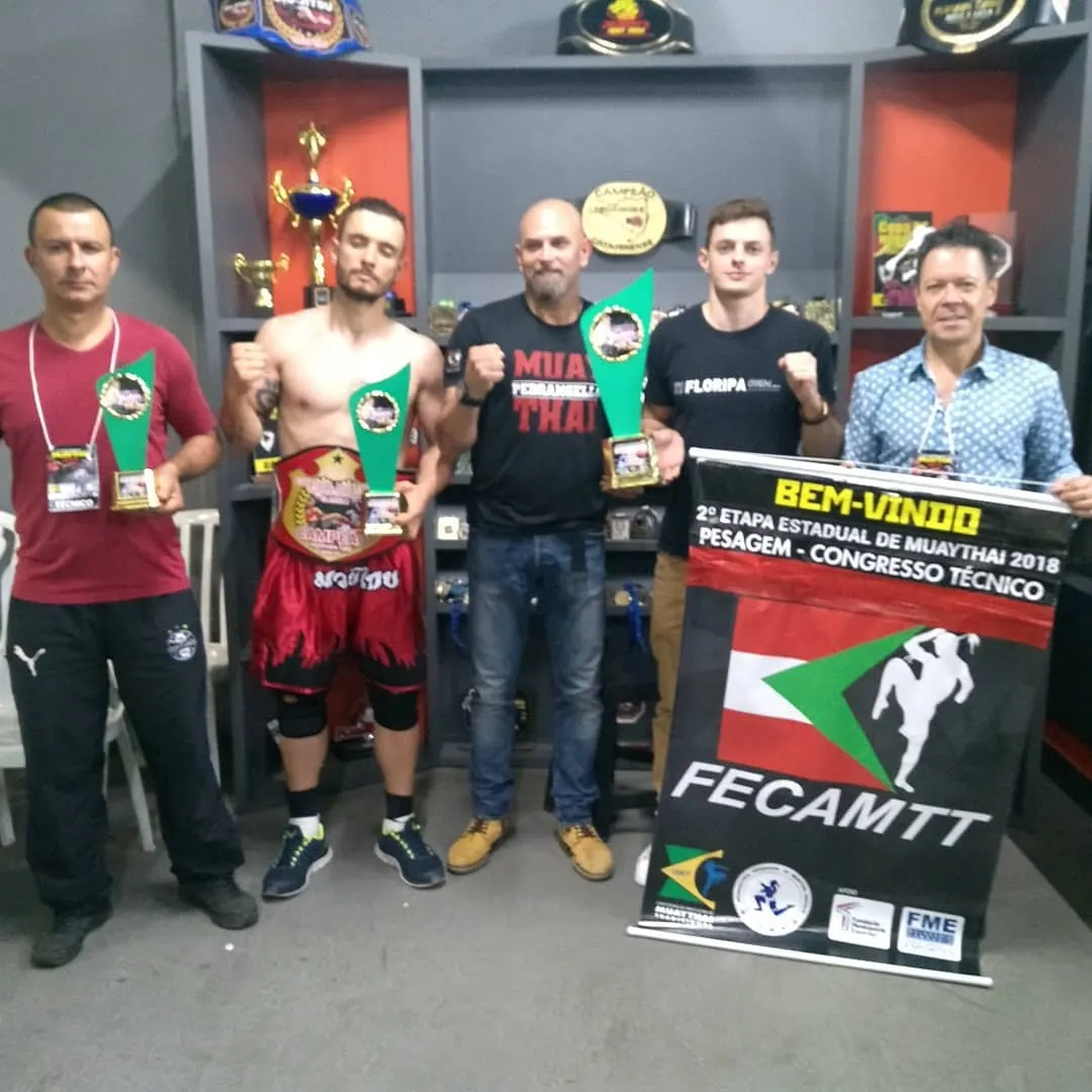 Mestre Paulo Ribeiro segurando o banner da FECAMTT da 2ª Etapa Estadual de Muaythai 2018, ao lado de atletas com troféus e cinturão