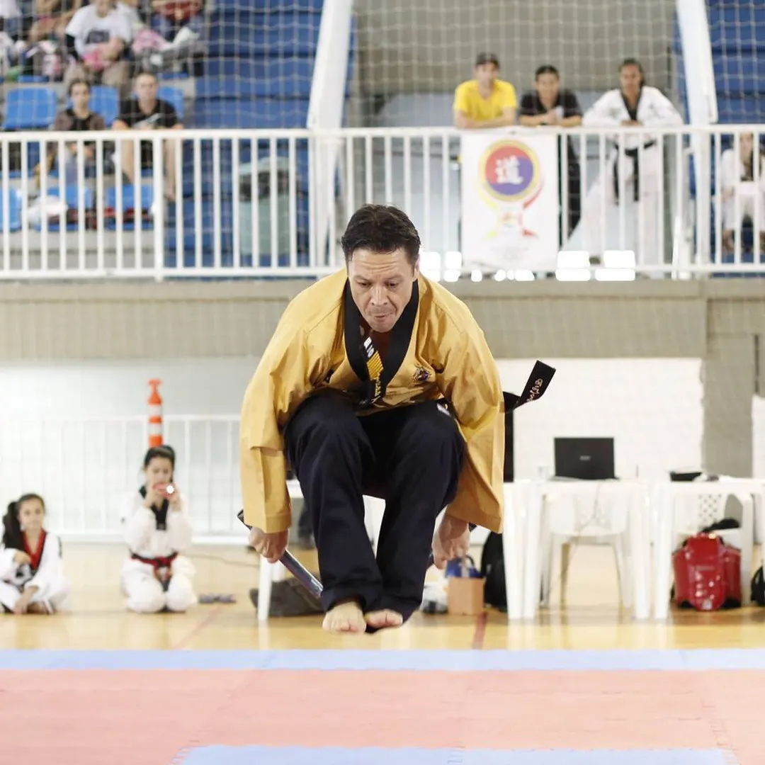 Mestre Paulo Ribeiro de dobok dourado saltando descalço sobre o tatame de um ginásio, com alunos de dobok assistindo ao fundo
