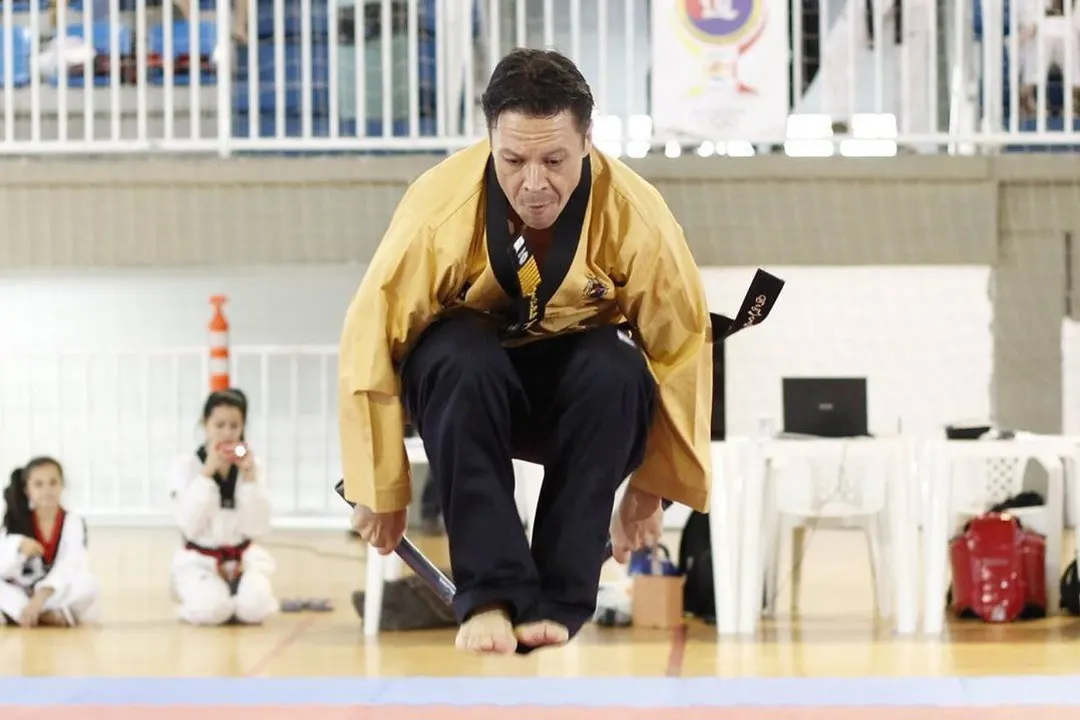 Mestre Paulo Ribeiro de dobok dourado saltando descalço sobre o tatame de um ginásio, com alunos de dobok assistindo ao fundo