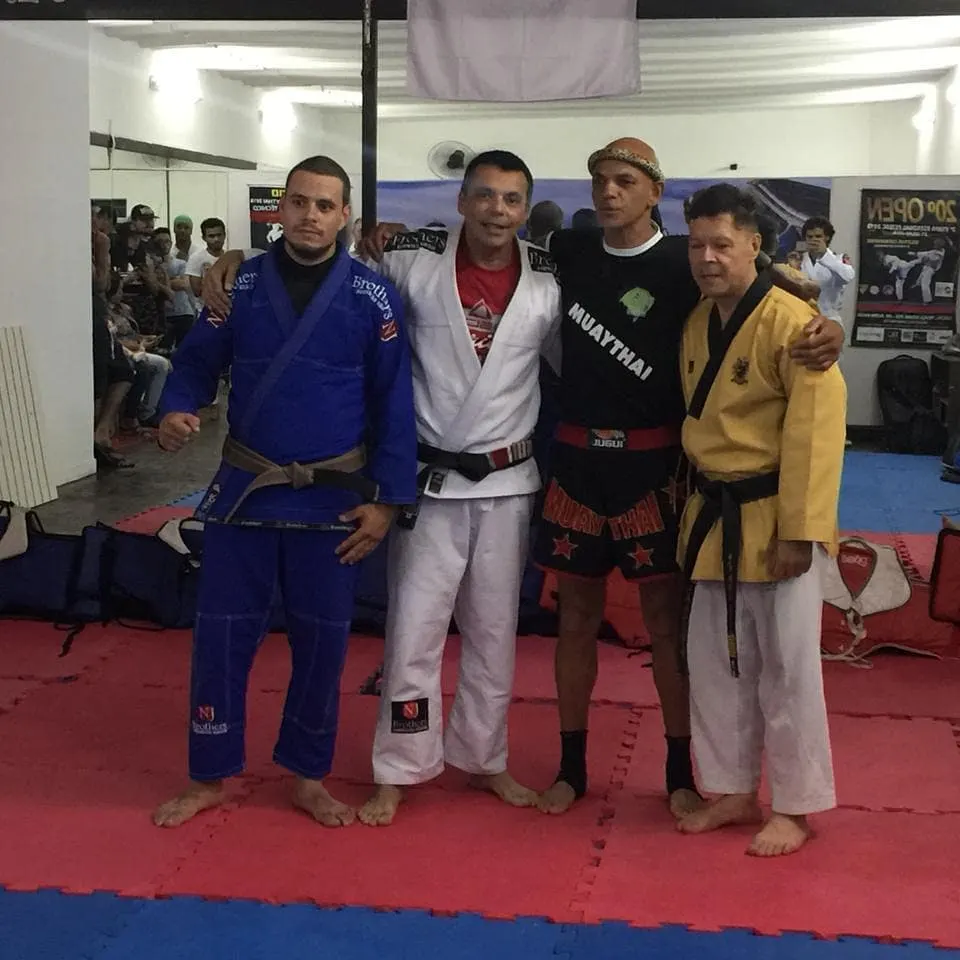 Mestre Paulo Ribeiro de dobok amarelo abraçado a três mestres convidados, de kimono azul, kimono branco e calção de Muaythai, sobre o tatame da academia, com público ao fundo