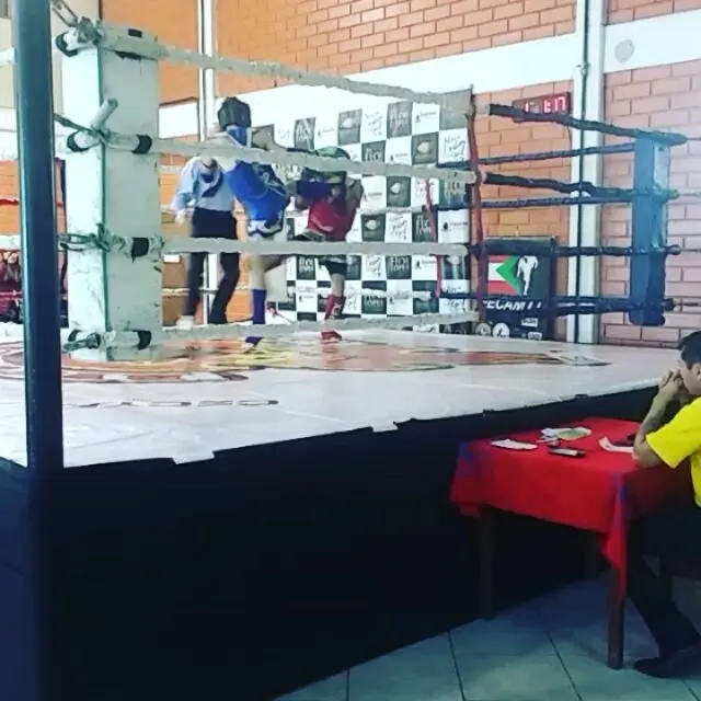 Luta de Muaythai num ringue montado em ginásio, com mesa de arbitragem e faixa da FECAMTT