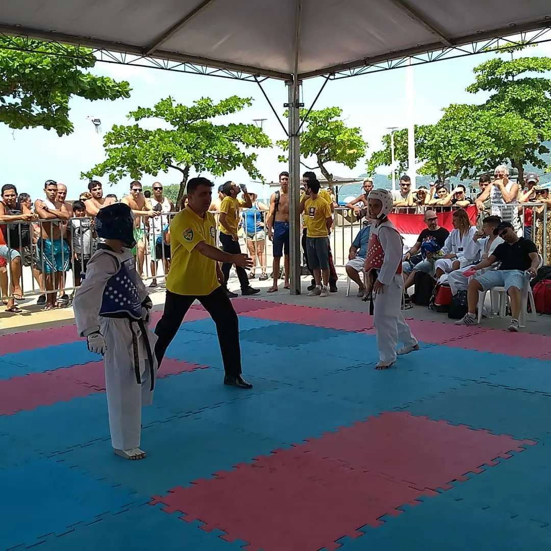 Luta infantil de Taekwondo sob tenda na orla, com árbitro e público assistindo atrás da grade