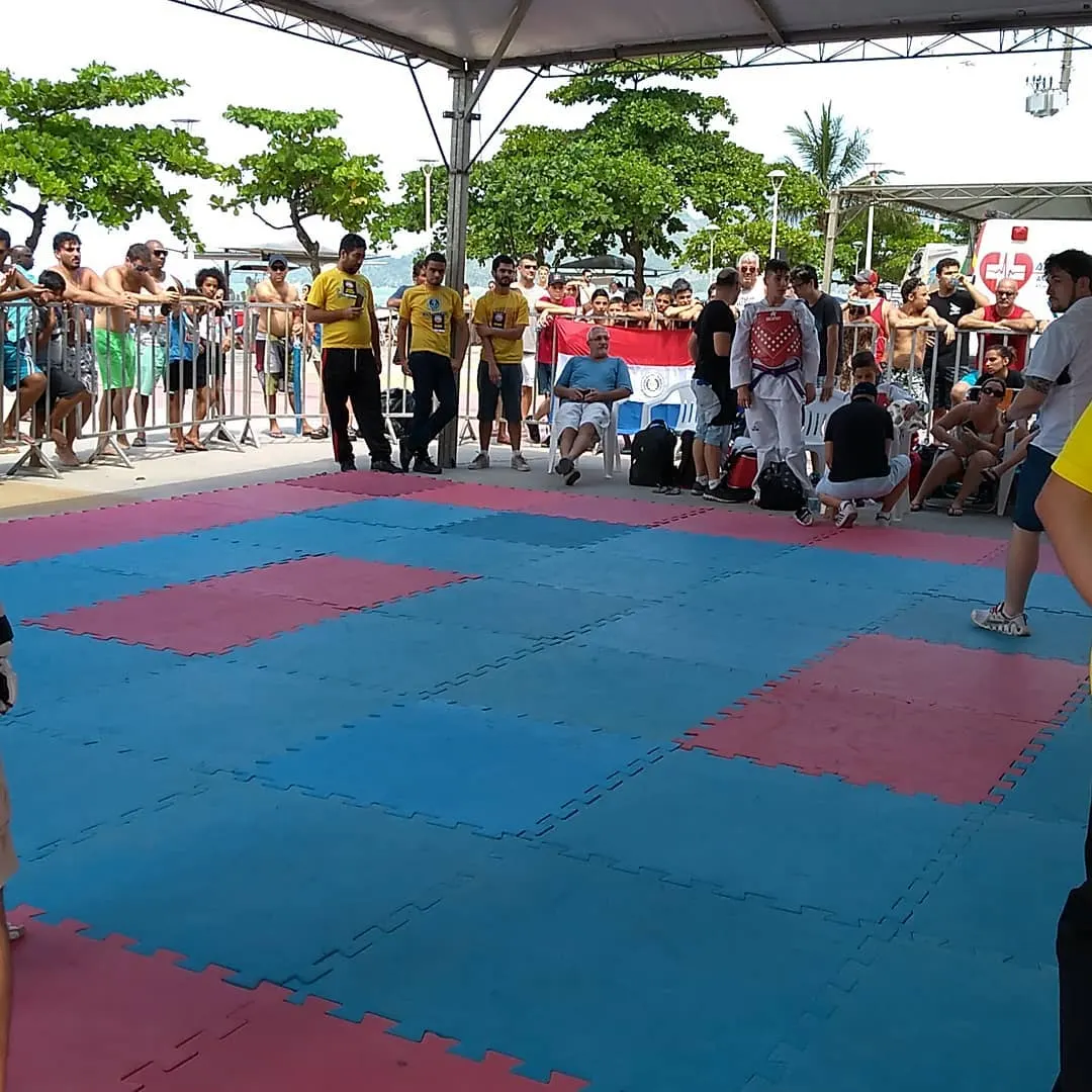 Tatame azul e vermelho sob tenda na orla, com árbitros de camisa amarela e público nas grades, uma bandeira do Paraguai ao fundo