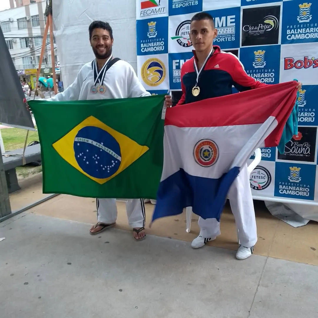 Dois medalhistas adultos segurando as bandeiras do Brasil e do Paraguai diante do painel de patrocinadores com a marca da FECAMTT