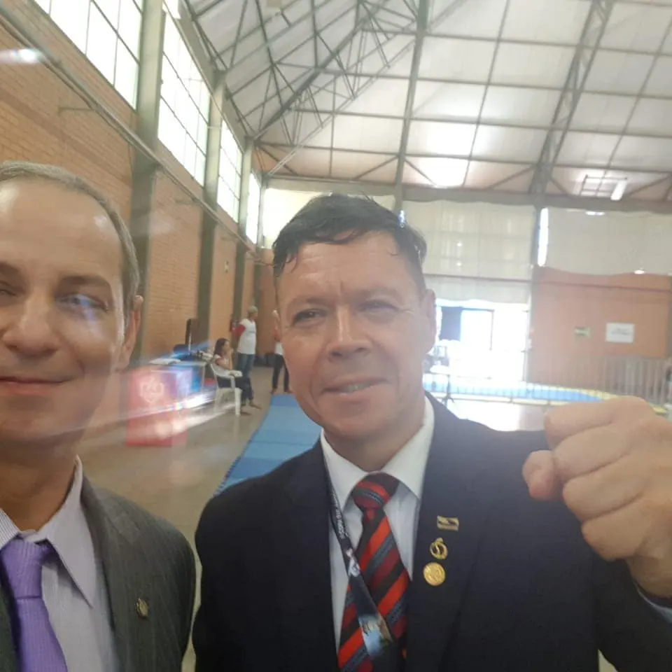 Mestre Paulo Ribeiro de terno e gravata, com o punho cerrado, ao lado do Mestre Alexandre num ginásio em Nova Petrópolis