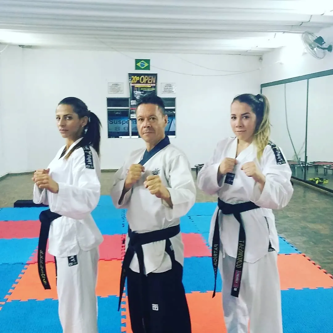 Mestre Paulo Ribeiro em posição de guarda entre duas alunas faixa preta, no tatame azul e vermelho da academia