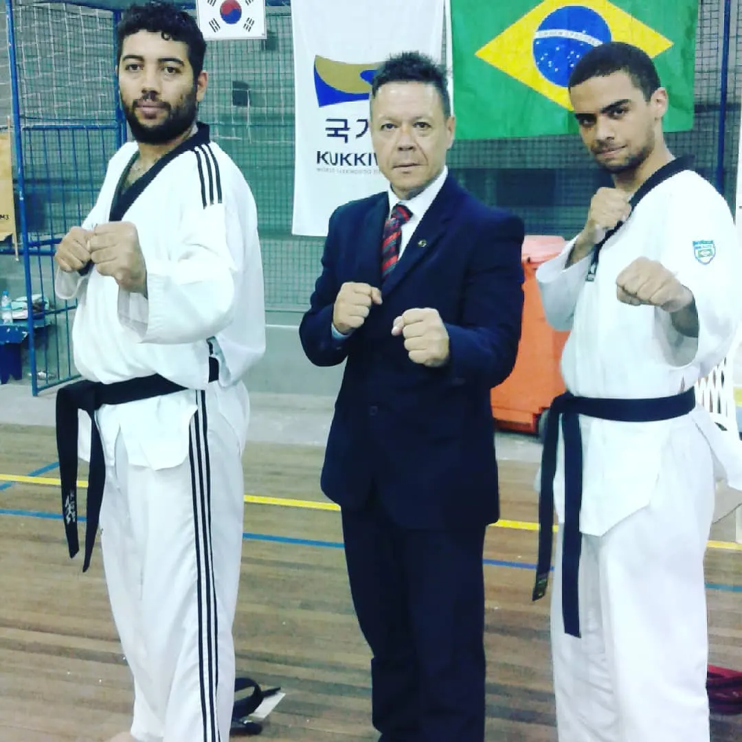 Mestre Paulo Ribeiro de terno, em guarda entre dois faixas-pretas, com as bandeiras da Coreia do Sul, do Kukkiwon e do Brasil ao fundo