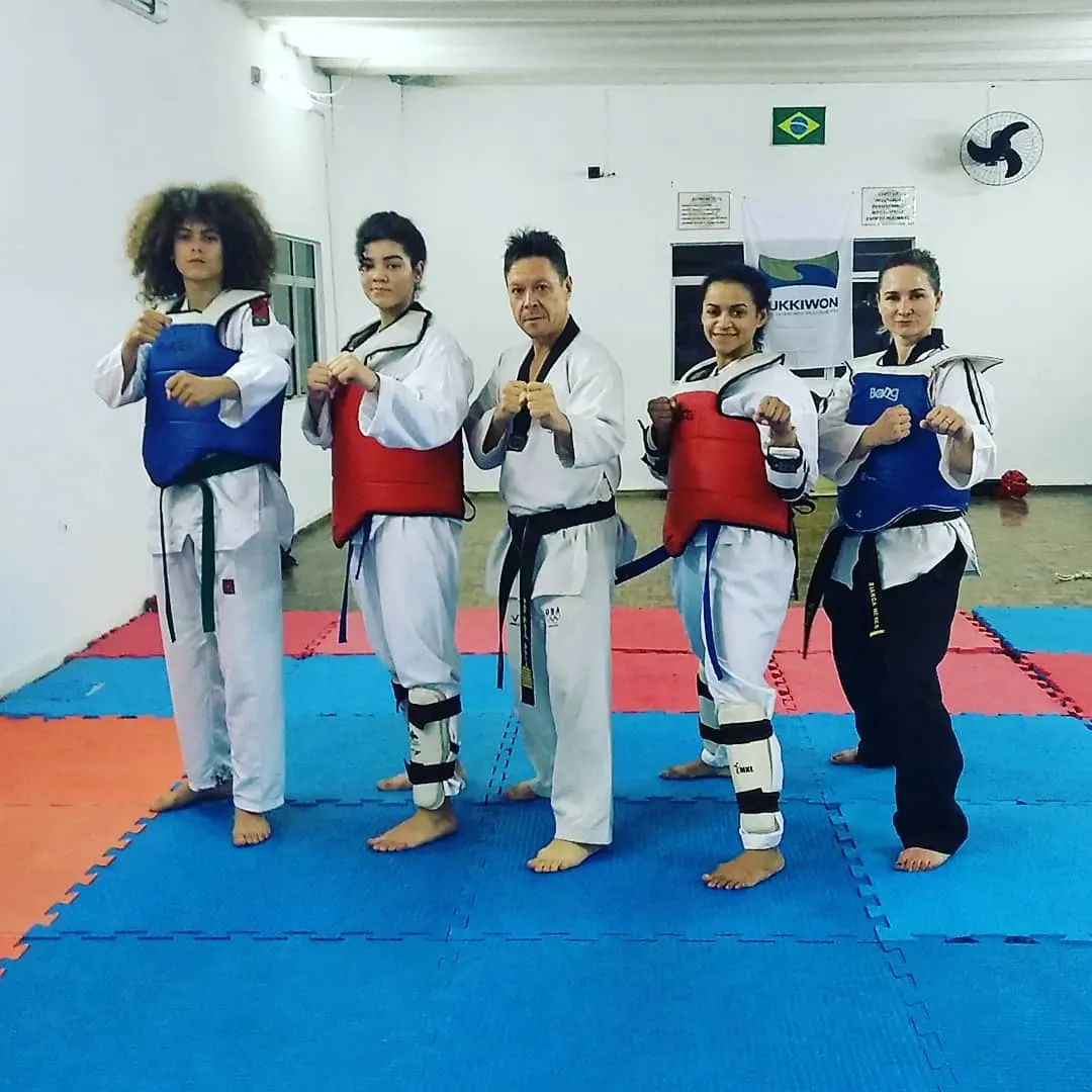 Mestre Paulo Ribeiro in a dobok and black belt, in fighting stance among four athletes in blue and red chest guards, in the academy with a Kukkiwon flag