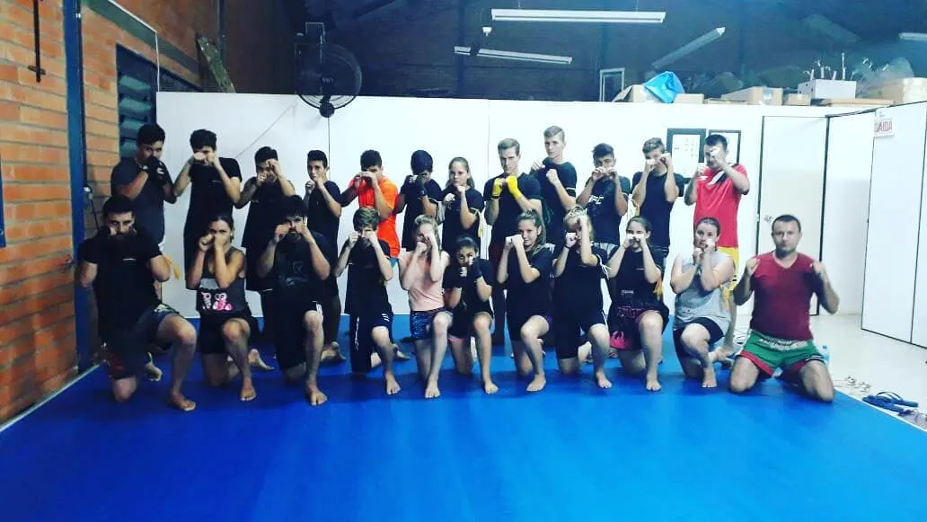 Equipe de Muaythai com cerca de vinte atletas em posição de guarda sobre o tatame azul