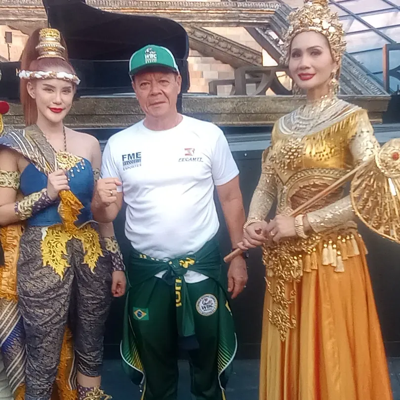 Mestre Paulo Ribeiro com camiseta da FECAMTT e boné da WBC Muaythai entre duas artistas com trajes tradicionais tailandeses