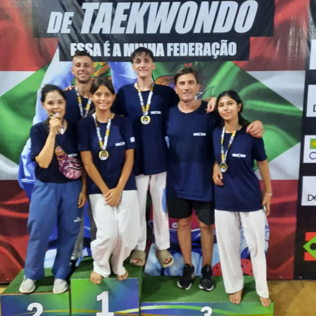 Atletas com medalhas no pódio, ao lado do técnico, diante de painel da federação de Taekwondo