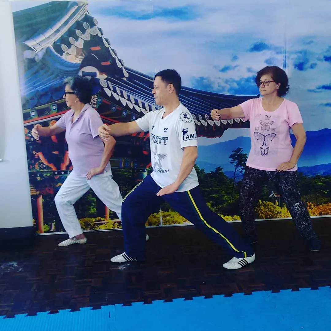Mestre Paulo Ribeiro conduzindo movimento de Tai Chi com duas alunas diante de um painel com templo coreano