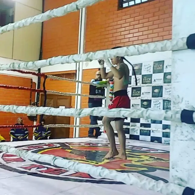 Atleta de calção vermelho fazendo o Wai Kru, a saudação tradicional do Muaythai, dentro do ringue
